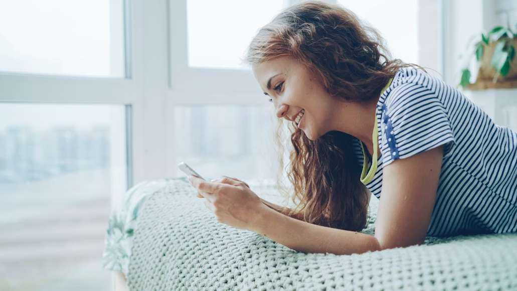 A happy young woman lying on her stomach on a bed and using a smartphone